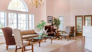 A spacious living room with a large window, a chandelier, a sofa, armchairs, and a coffee table. - Canberra Premiere Comfort Hotel (Mackinaw City)