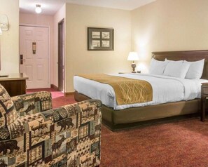 A hotel room with a bed, a chair, a nightstand, a lamp, and a door.