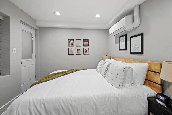 A bedroom with a bed, a nightstand, an air conditioning unit, and framed pictures on the wall.