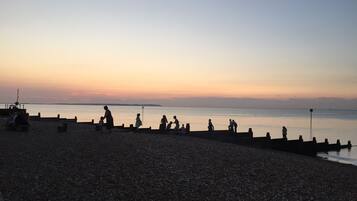 Une plage de galets où l’on voit des gens assis et debout le long du rivage, contemplant l’eau calme au coucher du soleil.