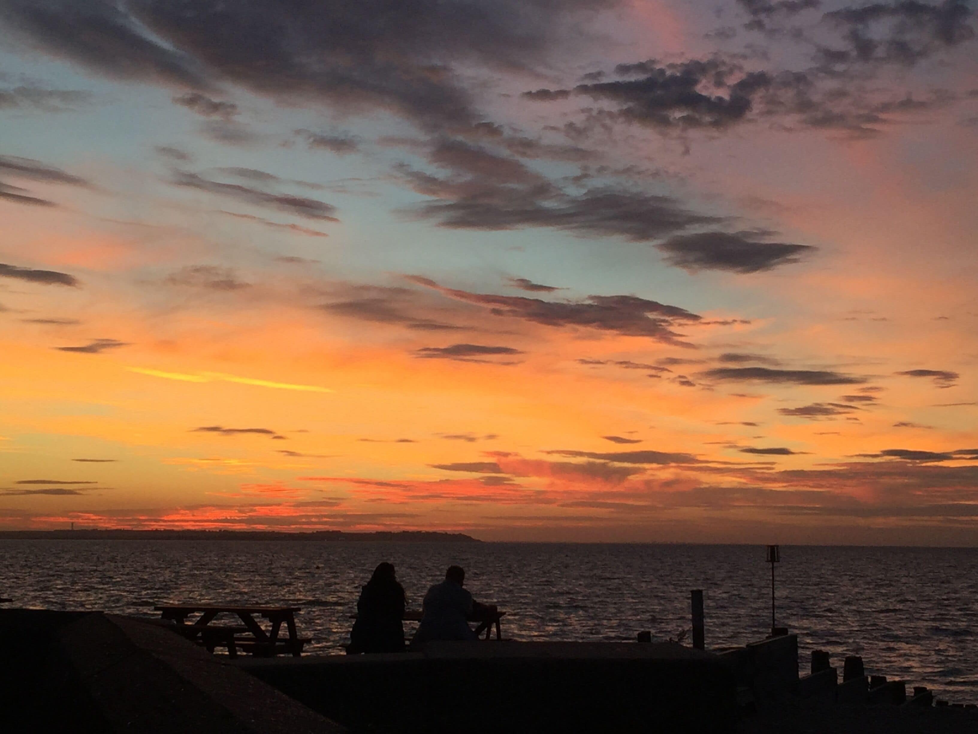 Un coucher de soleil sur l’océan, avec les silhouettes de personnes et d’un banc.
