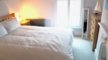 A neatly made bed with a white comforter, a wooden nightstand with a lamp, a dresser with a mirror, and a window with blinds.