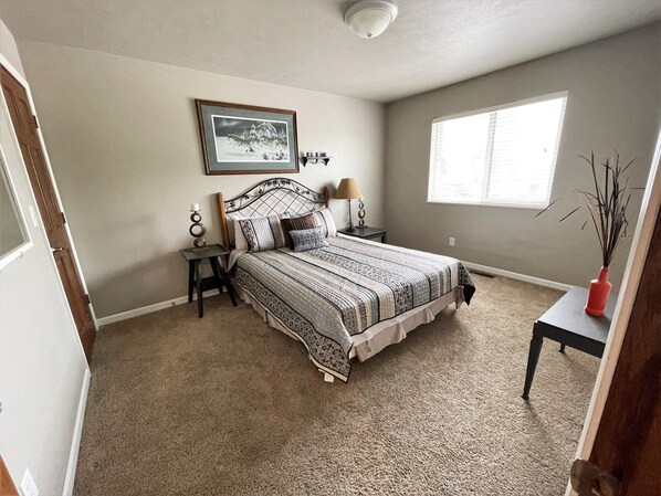 A bedroom with a large bed, a nightstand, a lamp, a framed picture on the wall, and a window.