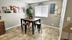 A dining area with a wooden table and two chairs, a potted plant, and wall decorations including a 'gather' sign and framed pictures. - Juniper Townhouse #4 (Mitchell)