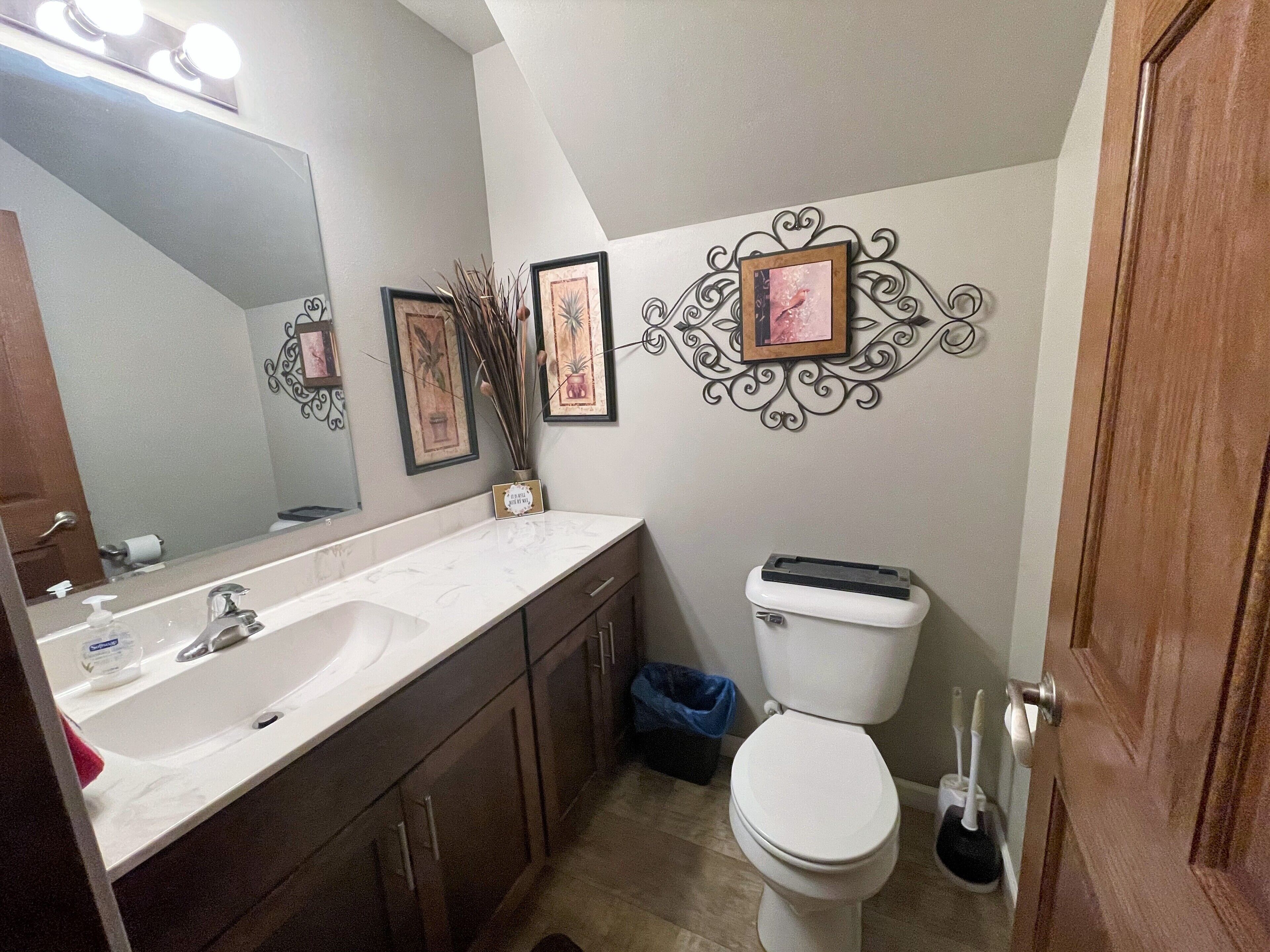 Een badkamer met een wit wastafel, spiegel en toilet. Er hangen afbeeldingen in lijsten aan de muur en een decoratief metalen object boven het toilet.
