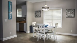 A dining area with a round table and white chairs, a kitchen in the background, and decorative wall art.