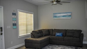 A living room with a sectional sofa, a ceiling fan, a beach-themed painting, and a window with blinds.