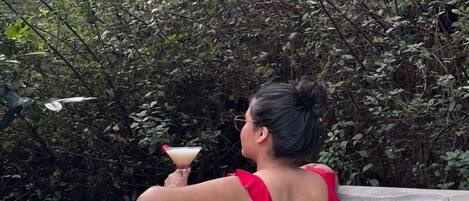 A person in a hot tub with a drink, surrounded by dense foliage.