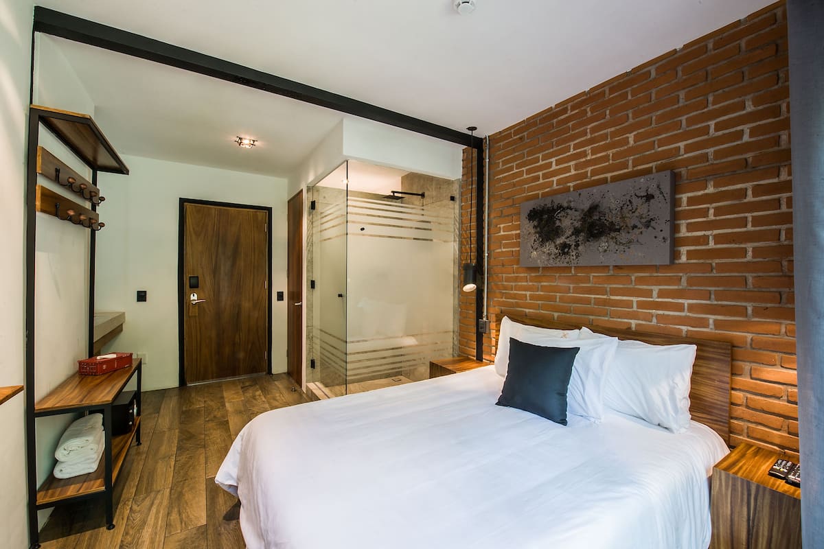 A modern bedroom with a large bed, a wooden nightstand, a glass shower enclosure, and a brick wall.
