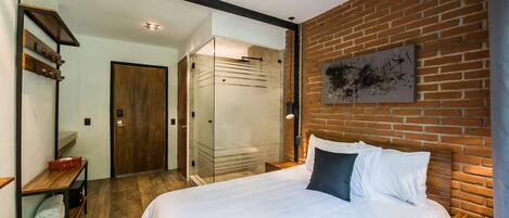 Une chambre moderne avec un grand lit, une table de chevet en bois, une cabine de douche en verre et un mur en briques.