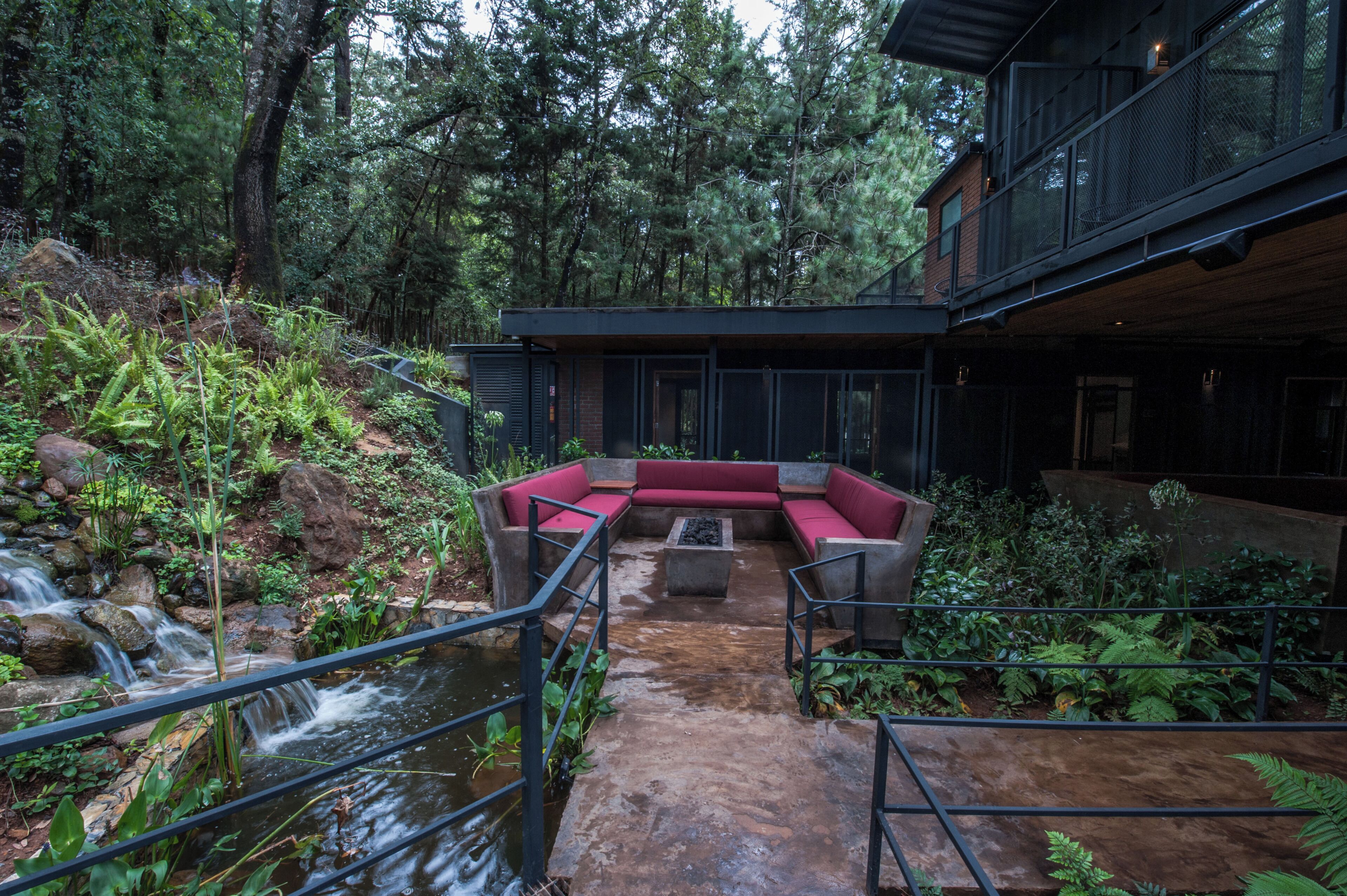 Un área de descanso moderna al aire libre con fuente de agua, rodeada de abundante vegetación y una terraza de madera.