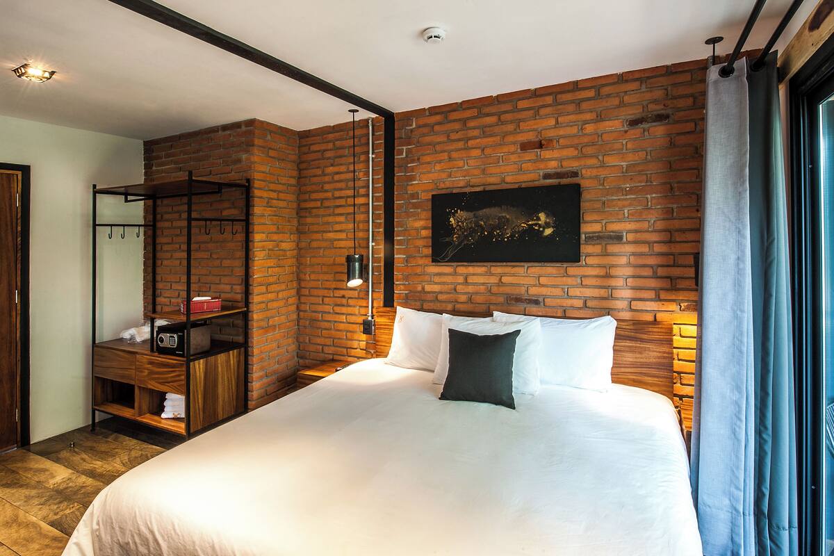 A hotel room with a brick wall, a bed with white bedding, a wooden nightstand, a coat rack, and a painting.