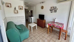 A compact living space with a green sofa, a small white table, a wooden TV stand, and a dining table with chairs. - 40m² With Terrace In A Residence With Pool (Le Grau-du-Roi)