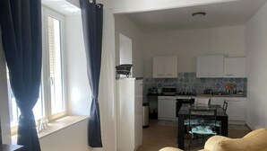 Una sala de estar con un sofá color beige, una mesita y un ventanal con cortinas. Se ve una cocina al fondo con armarios blancos y un salpicadero de azulejos.