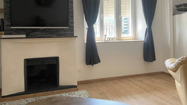 Una sala de estar con chimenea de piedra, un televisor de pantalla plana montado encima y una ventana con cortinas, además de un sofá color beige.