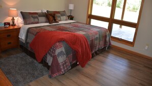 A bedroom with a large bed, a nightstand with a lamp, a window with a view, and a wooden floor.