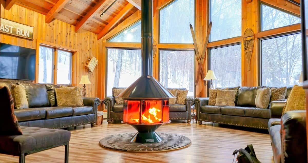 Un salon chaleureux avec deux canapés en cuir, un poêle à bois central et de grandes fenêtres donnant sur la forêt.