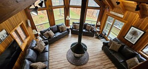 A cozy cabin interior with a central wood-burning stove, leather sofas arranged around it, and large windows offering views of the outdoors.