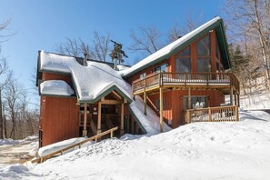 A cabin with a steep roof, a wooden deck, and a staircase leading to a second-story balcony.