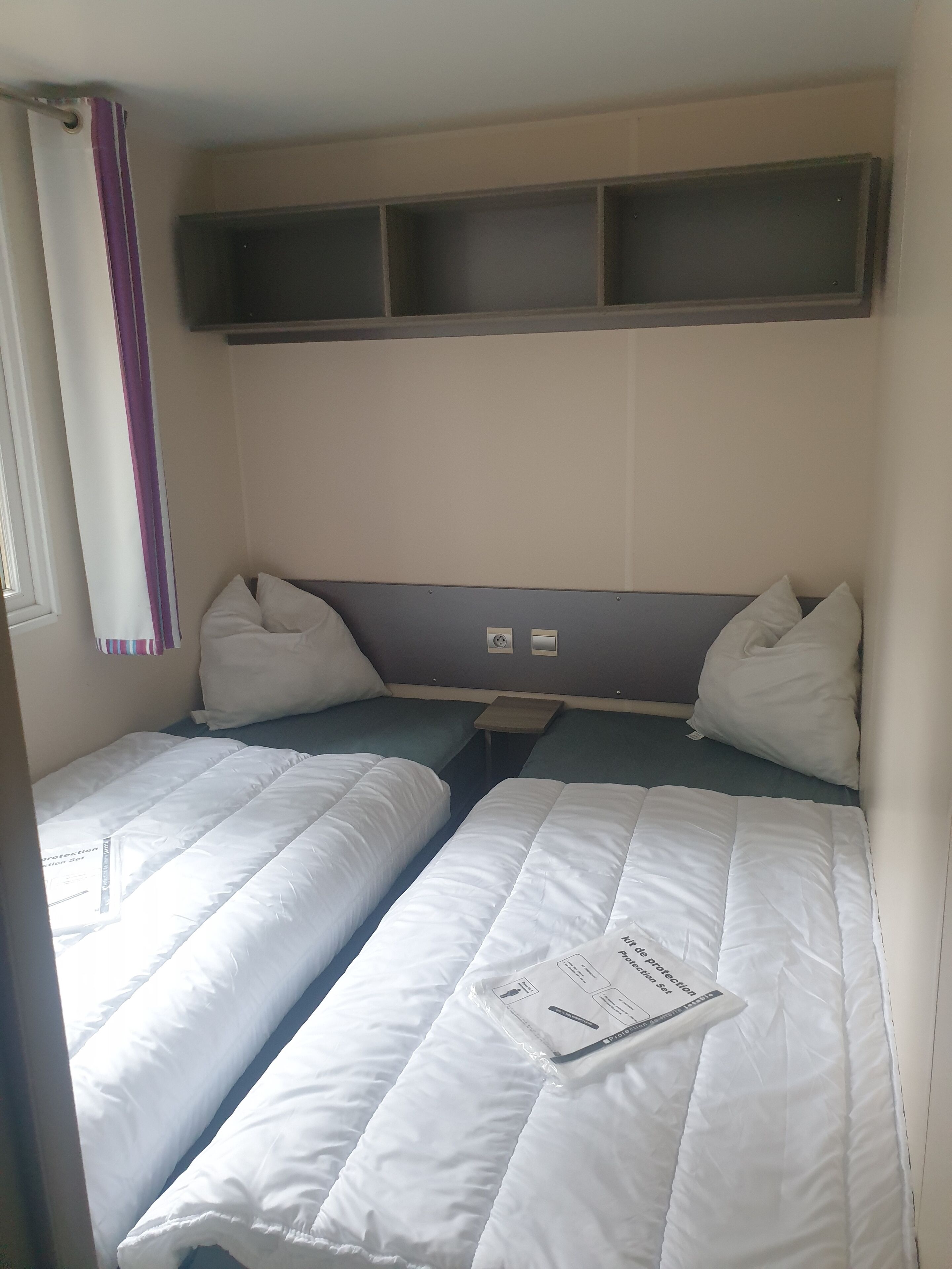 Une chambre avec deux lits simples, chacun équipé de draps et d’oreillers blancs. On trouve deux prises électriques sur le mur, au-dessus des lits.