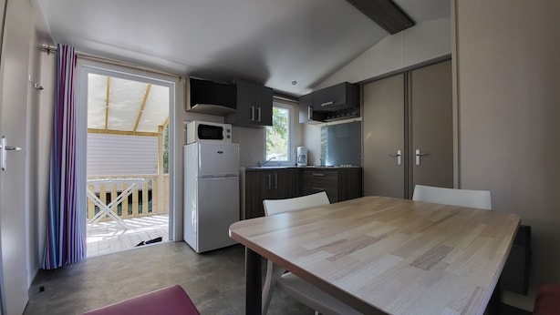 Une cuisine compacte équipée d’un réfrigérateur, d’un four à micro-ondes et de placards. On y trouve une table à manger avec des chaises, ainsi qu’une porte-fenêtre coulissante donnant sur un espace extérieur.