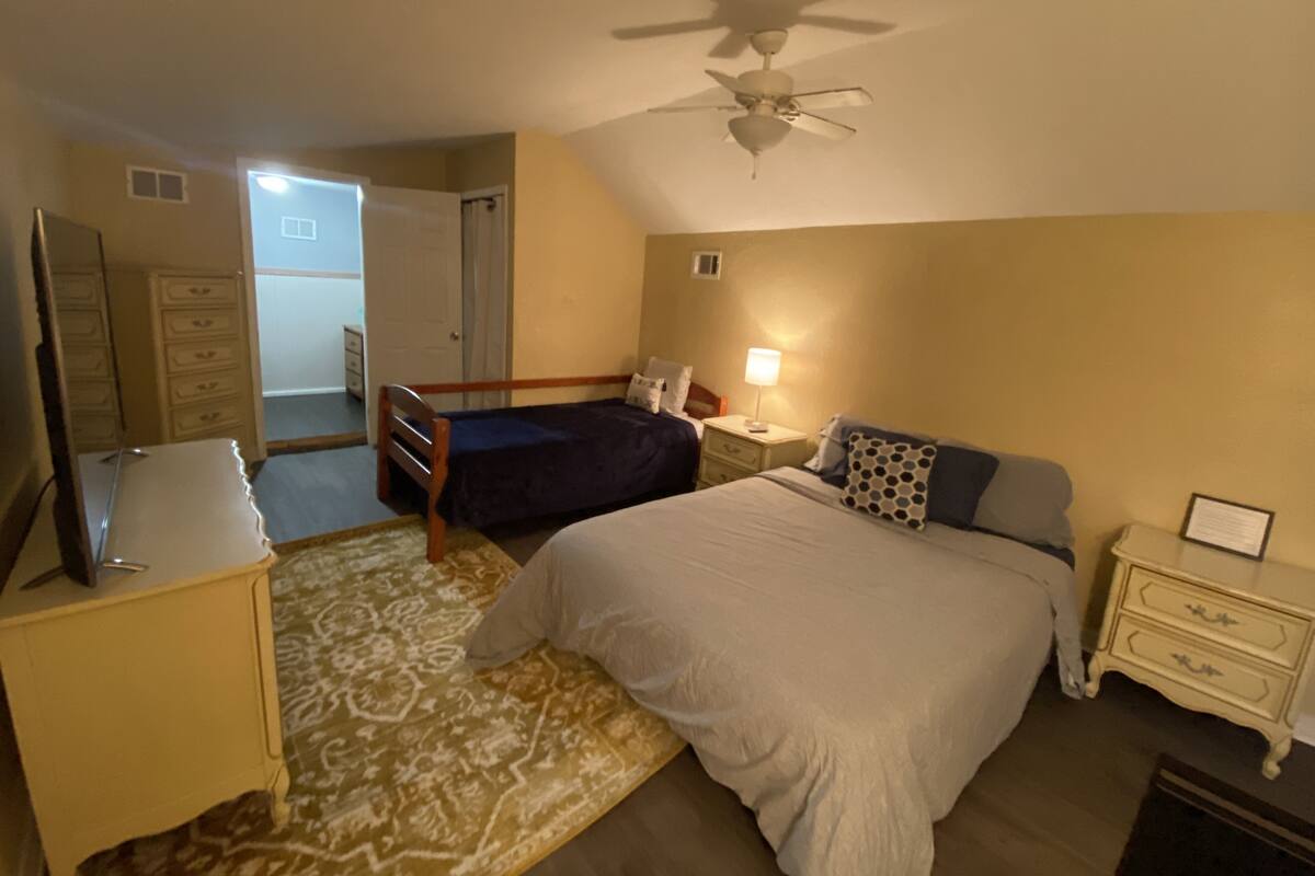 A bedroom with a bed, bedside tables, a desk, a chair, a ceiling fan, and a door leading to another room.