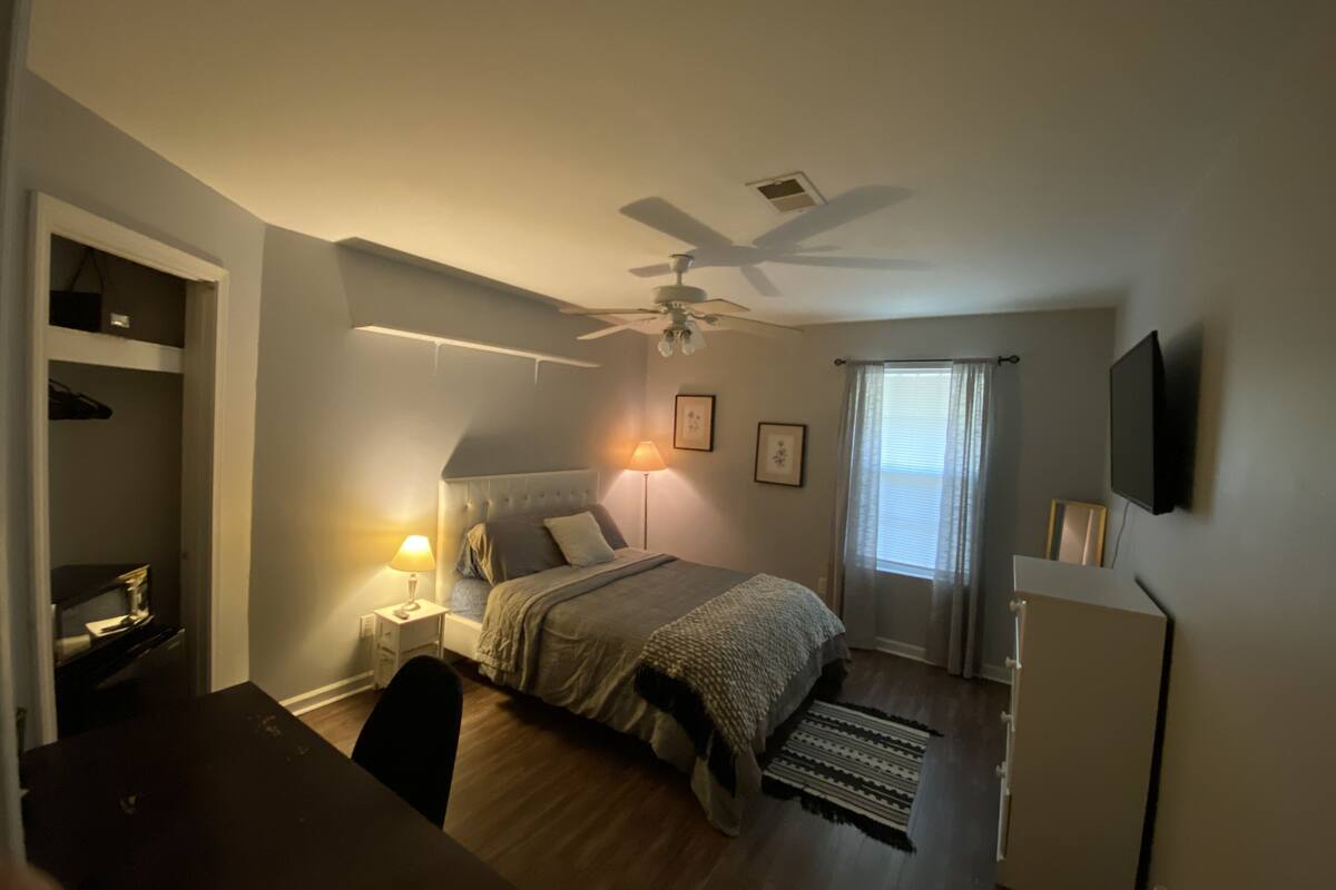 A bedroom with a bed, bedside lamps, a ceiling fan, a TV, and a window with curtains.