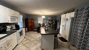 Una cocina con armarios blancos, microondas, estufa y refrigerador. Hay una mesa de comedor con sillas y una cortina estampada.
