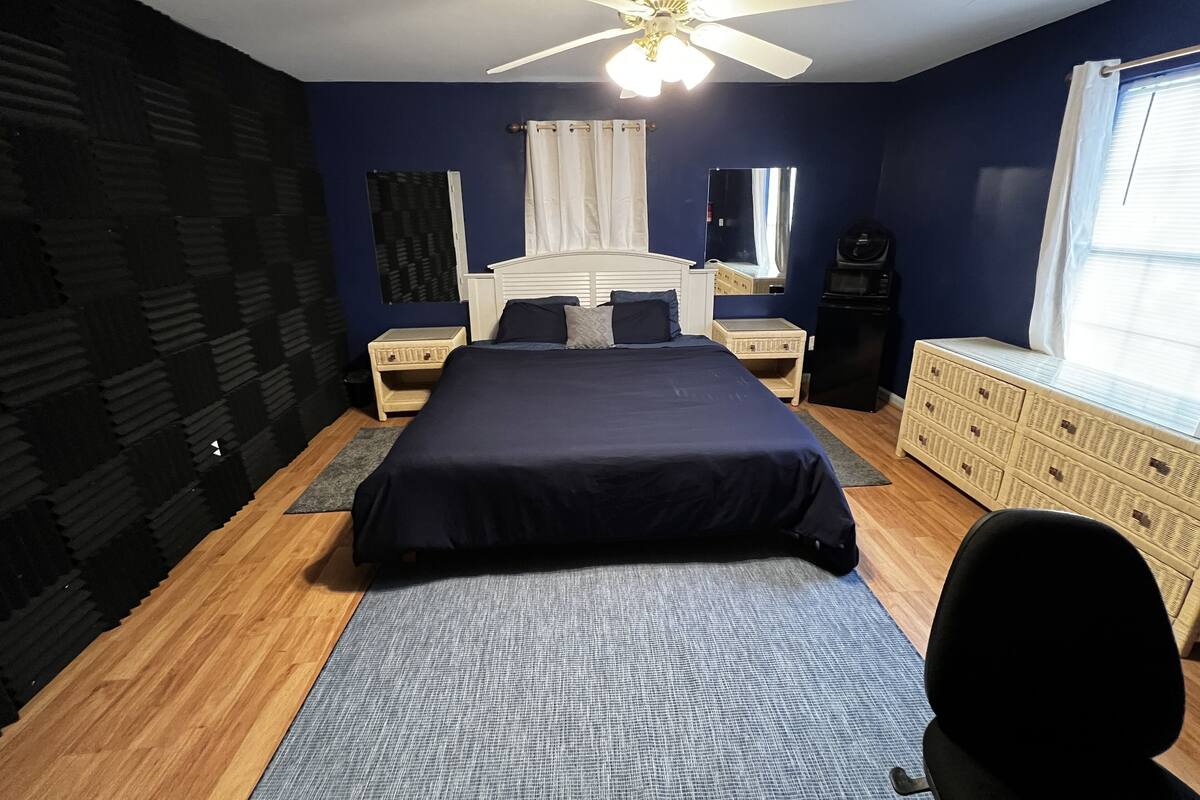A bedroom with a bed, a ceiling fan, a dresser, a window with curtains, and a microwave.