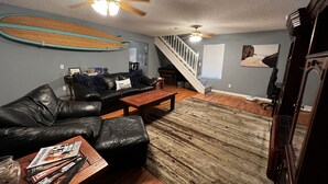 Una sala con un sofá de cuero, una mesa de centro de madera y una alfombra. Se ve una escalera que sube a otro nivel, y un tabla de surf está colgada en la pared.