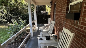 Un patio techado con mecedoras blancas y una mesa, una pared de ladrillo y una ventana.