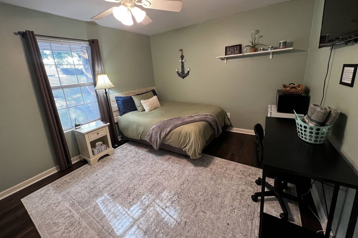 A bedroom with a bed, a desk, a chair, a ceiling fan, a window with curtains, and a wall-mounted shelf with decorative items.