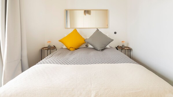 A neatly made bed with a quilted grey blanket and two pillows, one yellow and one grey, in a simple room with a mirror on the wall and two bedside tables with lamps.
