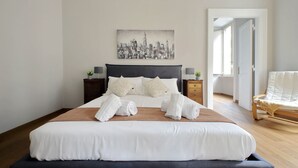 Een net opgemaakt bed met witte lakens, een stadsgezichtschilderij erboven en een houten vloer.