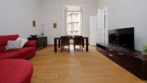 Een woonkamer met een rode bank, een eettafel met stoelen, een televisie op een kast en uitzicht op een gebouw door een geopende deur.