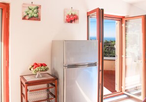 Uma cozinha com geladeira, uma fruteira sobre um suporte de madeira e uma porta de vidro deslizante que dá para uma varanda com vista para o mar.