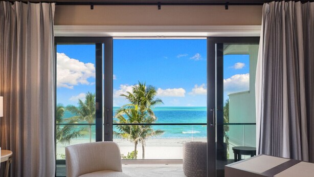 A modern room with a large sliding glass door leading to a balcony with a view of the beach and ocean.