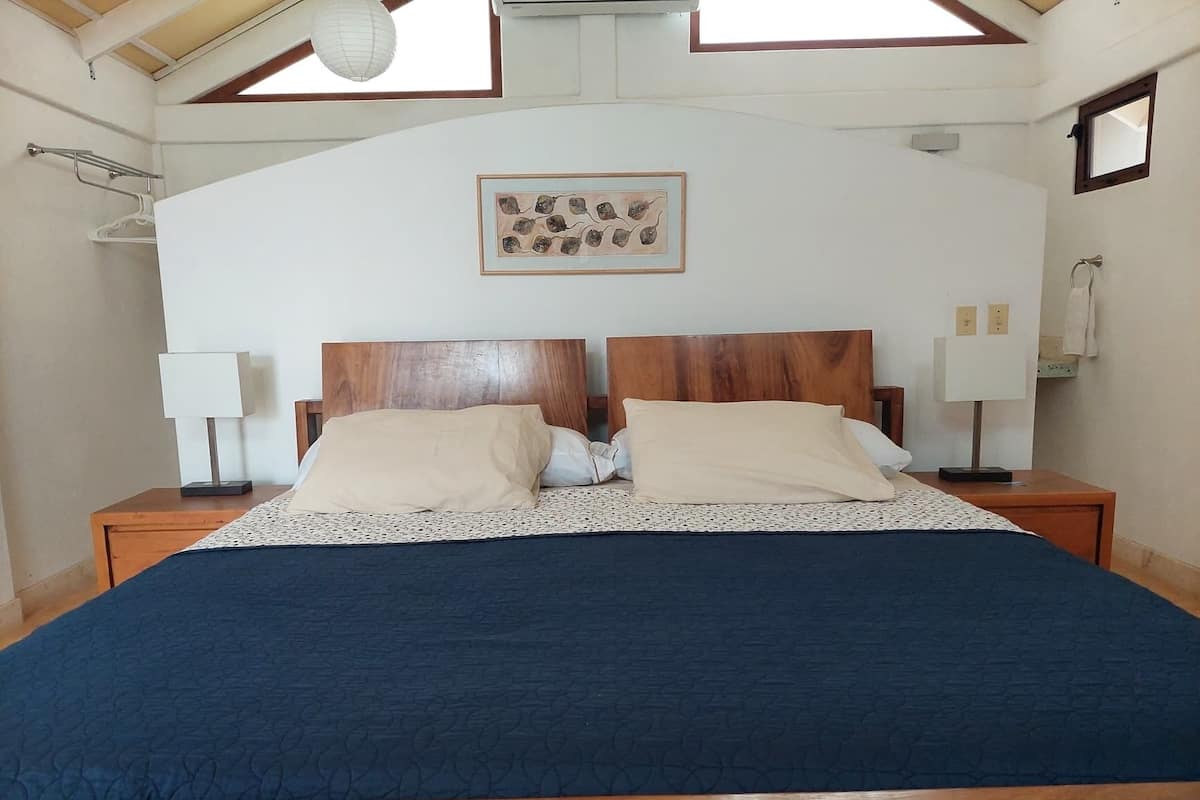 Un dormitorio con cama de madera, dos mesitas de noche con lámparas, un cuadro enmarcado en la pared y un tragaluz arriba.