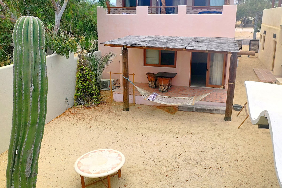 Un pequeño patio al aire libre con una mesa y sillas de madera, una hamaca y un cactus.