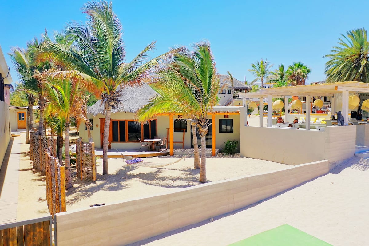 Un resort frente a la playa con palmeras, un edificio de techo de paja y una zona de estar techada.