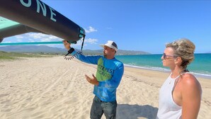 Una persona sostiene una tabla de surf en una playa, con cielos despejados y montañas al fondo.