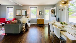 A modern living room with a grey sofa, red chairs, a wooden herringbone floor, a dining area with white chairs and a table, and a large window with a view of greenery. - Walk to St Charles Main St. Shops, Eateries & Bars! (Saint Charles)