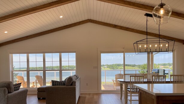 A spacious room with a high ceiling, wooden beams, a large window overlooking a body of water, a cozy seating area with a sofa and chairs, and a dining area with a table and chairs. - Lady Bug Lane Escape, Clinton, PEI (Clinton)