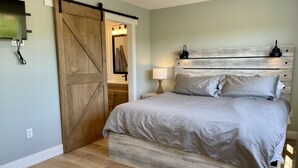 A bedroom with a large wooden sliding door, a bed with light-colored bedding, a bedside table with a lamp, and a headboard with light fixtures. - Lady Bug Lane Escape, Clinton, PEI (Clinton)