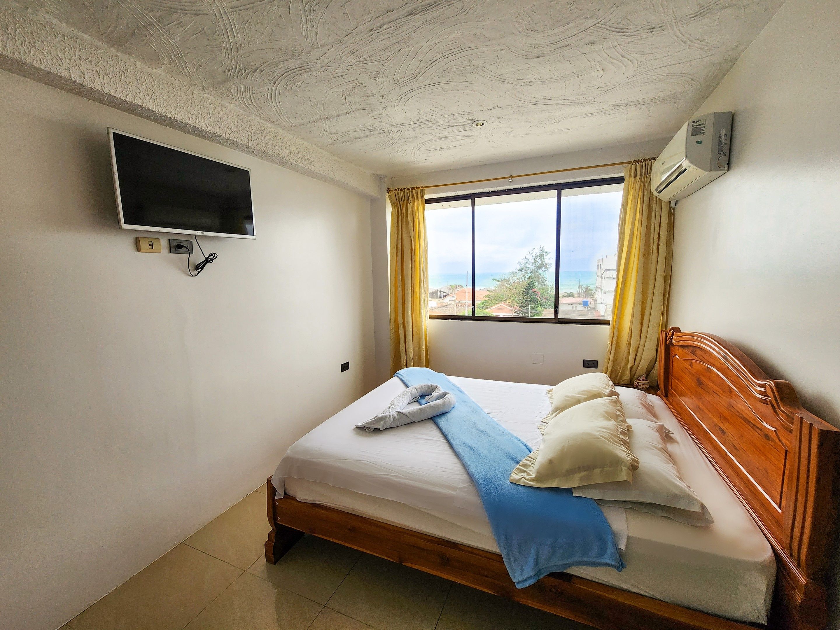 Een hotelkamer met een bed, een aan de muur bevestigde televisie, een airconditioning en een raam met gordijnen.