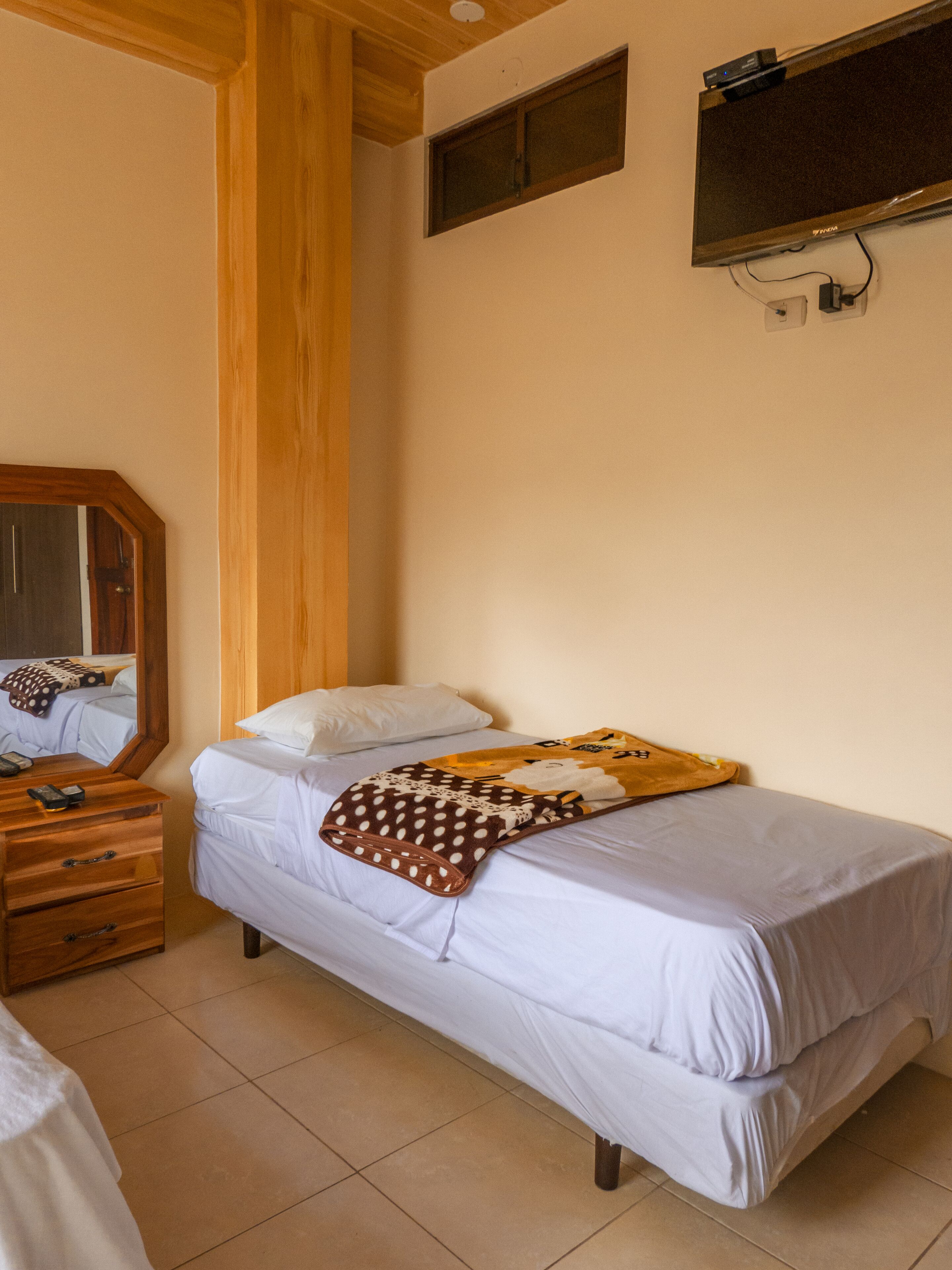 Un dormitorio con cama, cómoda de madera, espejo y televisión montada en la pared.