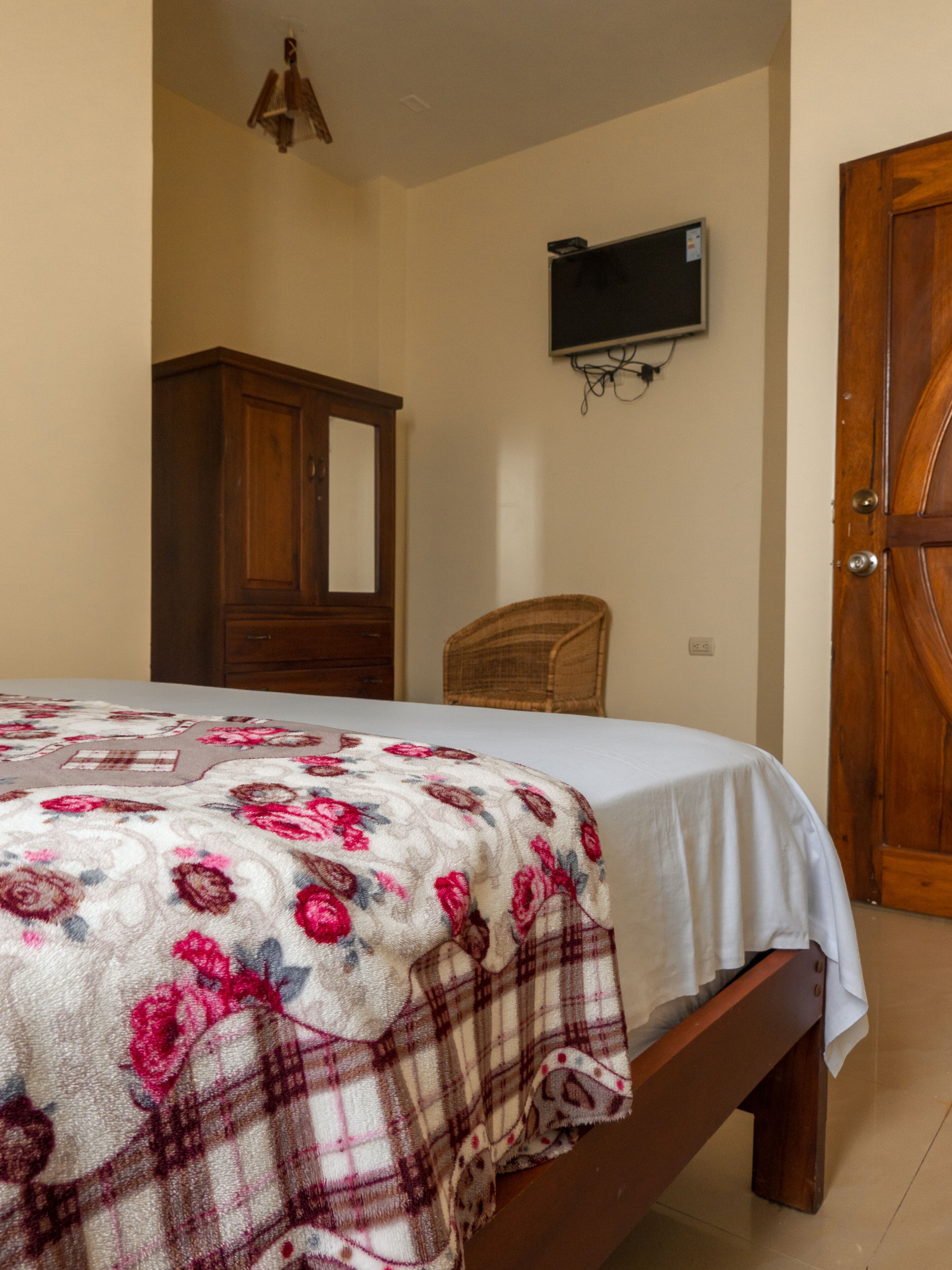A bedroom with a bed, a wooden wardrobe, a television mounted on the wall, and a wooden door.