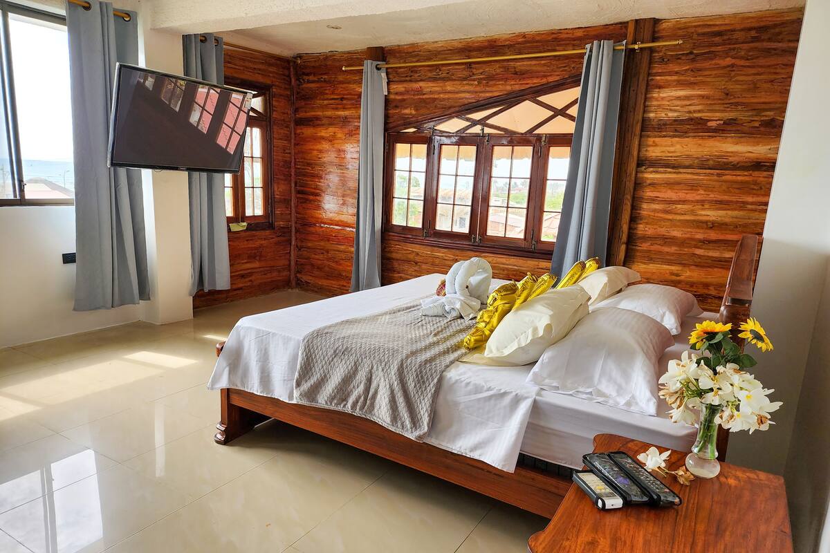 A bedroom with a wooden headboard, a bed with white and yellow bedding, a TV mounted on the wall, and a wooden nightstand with a vase of flowers.