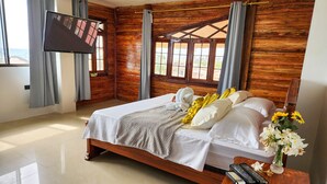 A bedroom with a wooden headboard, a bed with white and yellow bedding, a TV mounted on the wall, and a wooden nightstand with a vase of flowers. - Hostal Poseidon (Puerto Cayo)