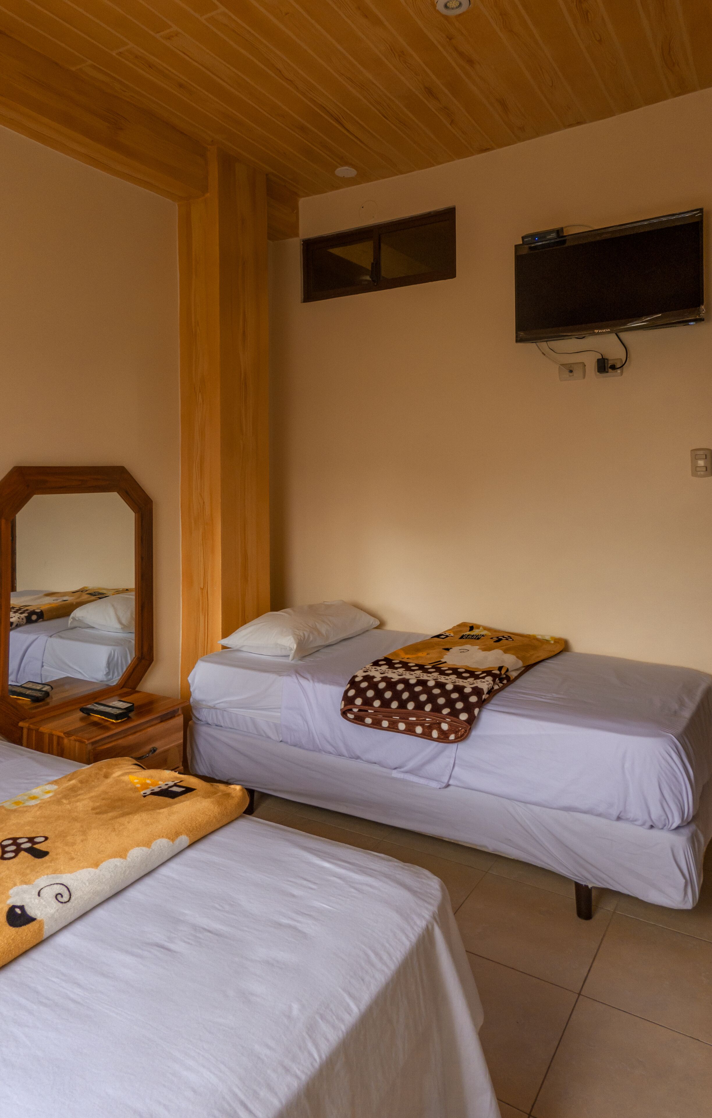 A room with two beds, a wooden ceiling, a mounted TV, and a mirror.